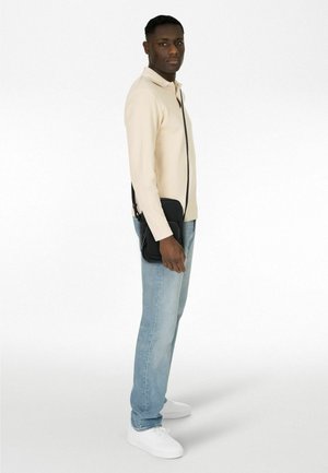 Borsa a tracolla nera con finitura texturizzata, indossata sopra un pullover beige, abbinata a jeans azzurri e sneakers bianche.