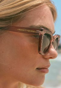 Kandilised prillid selgete, tekstuursete raamidega, roosa lisandiga ja kuldsete detailidega tempel. Läätsed on tumedad, pakkudes varju.