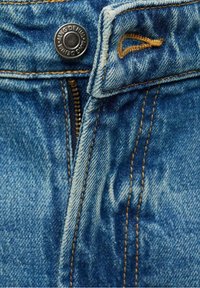 Gros plan d'un jean en denim bleu montrant une fermeture éclair partiellement ouverte, un bouton métallique avec le texte "KING DENIM" et des coutures jaunes visibles.