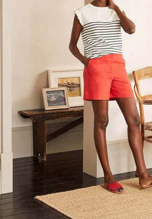 Rote Shorts und ein weißes Oberteil mit marineblauen Streifen, versehen mit gerafften Ärmeln. Das Umfeld besteht aus Holzmöbeln und einem gewebten Teppich.