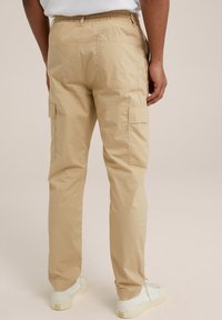 Man som bär beige cargobyxor och vita sneakers, står vänd bort mot en enkel ljus bakgrund.