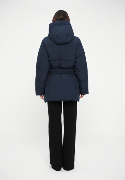 Doudoune marine avec taille ceinturée, capuche et poignets élastiques. Texture lisse avec une coupe mi-longue, portée sur un pantalon large noir.