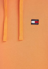 Oranje trui met een gladde textuur, voorzien van twee trekkoorden en een klein rechthoekig label met "TOMMY JEANS" in blauw, wit en rood.