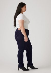 Mörkblå denimjeans med smal passform, utrustade med bakfickor och en läderlapp. Kombinerade med en figurnära vit t-shirt och svarta högklackade skor.