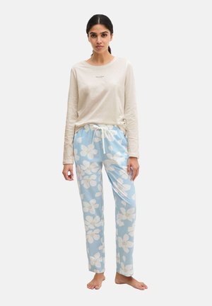 Vrouw staat blootsvoets, draagt een crèmekleurige top met lange mouwen en lichtblauwe broek met trekkoord en grote witte bloemmotieven.