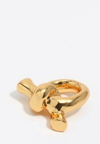 Anillo dorado brillante con tres protuberancias redondeadas, colocado sobre una superficie blanca.