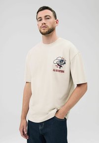 T-shirt en coton beige clair à manches courtes, arborant un design graphique d'un crâne portant un chapeau de cow-boy et le texte "TOUT OU RIEN" en rouge.
