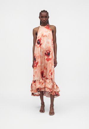 Nő ujjatlan őszibarack és piros virágmintás pántos ruhában, fodros szegéllyel és pántos magas sarkú szandálban, egyszínű háttér előtt áll.