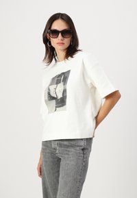 Tricou alb din bumbac cu croială oversized, având un imprimeu grafic alb-negru pe față. Modelul poartă blugi gri și ochelari de soare.