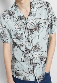 Camicia a maniche corte in azzurro chiaro con stampa di ananas neri. Presenta una tasca, chiusura frontale con bottoni e collo classico.