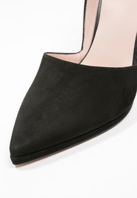 Chaussure à talon haut en daim noir avec un bout pointu et un design découpé. L'intérieur présente un revêtement lisse rose clair et un talon élancé.