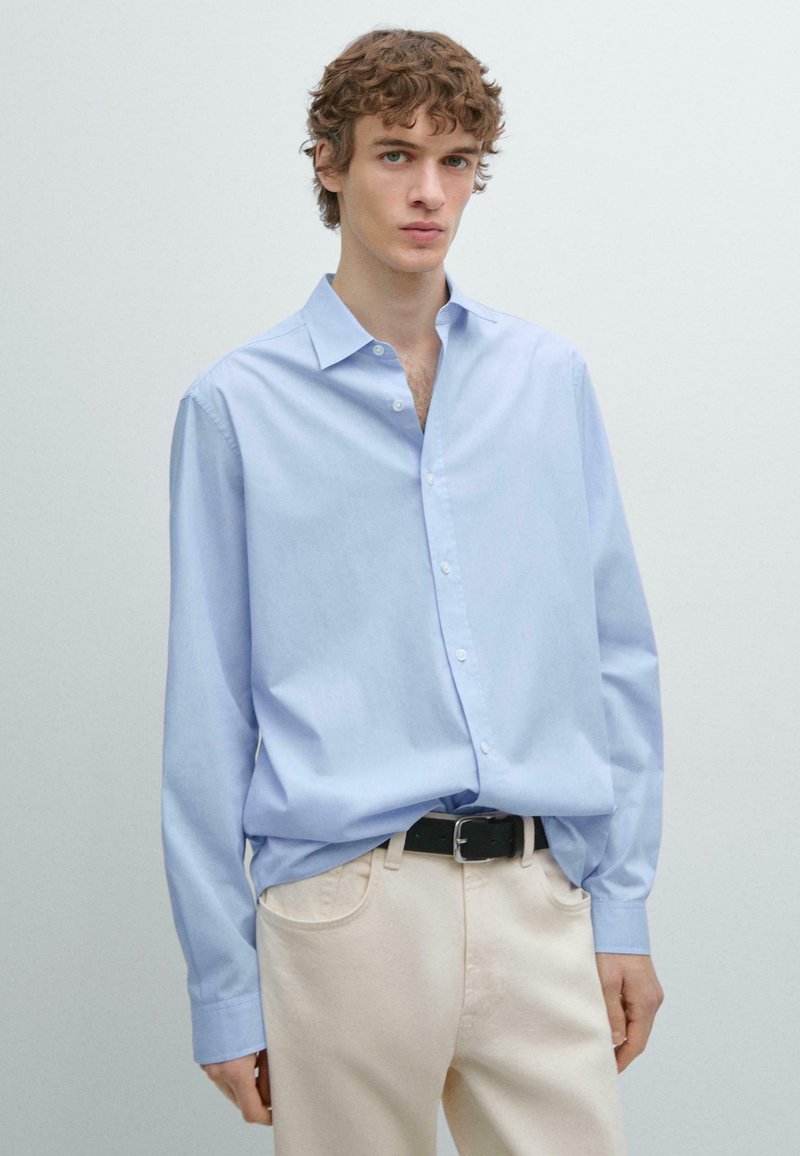 Světle modrá košile na knoflíky z měkké bavlny, s klasickým límcem a dlouhými rukávy, nošená s krémovými kalhotami a černým opaskem.