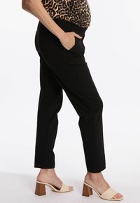 Pantalons noirs ajustés avec poches, fabriqués à partir d'un tissu texturé. Portés avec des sandales à talons carrés beige. Profil latéral visible.