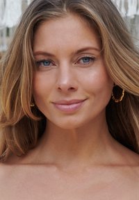 Boucles d'oreilles en or avec un design tordu, cheveux châtains clairs avec des reflets subtils, peau lisse et yeux bleus sur un fond neutre.