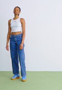 Balta trumpa tankų palaidinė, mėlyni laisvi džinsai ir šviesiai mėlyni sportbačiai su mediniu padu, prieš neutralų foną.