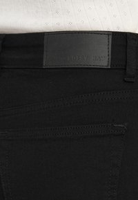 Κοντινό πλάνο μαύρων τζιν από τζιν με πίσω τσέπη και μαύρη δερμάτινη ετικέτα με την επιγραφή "NOISY MAY" κάτω από ένα λευκό πλεκτό τοπ.