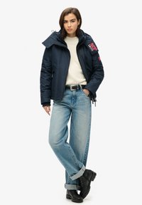 Granatowa kurtka puchowa z wysokim kołnierzem, zamkiem z przodu i detalem z logo na rękawie; w połączeniu z jasnoniebieskimi spodniami typu straight-leg i czarnymi botkami.