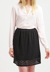 Helle rosa Langarmbluse mit V-Ausschnitt und Knopfdetails, kombiniert mit einem schwarzen Rock mit geometrischem Ausschnittmuster.