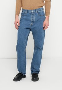 Ανοιχτό μπλε τζιν παντελόνι με χαλαρή εφαρμογή, που διαθέτει ζώνη μέσης με κούμπωμα κουμπιού και αντίθετη ραφή. Συνδυάζεται με μαύρα παπούτσια.