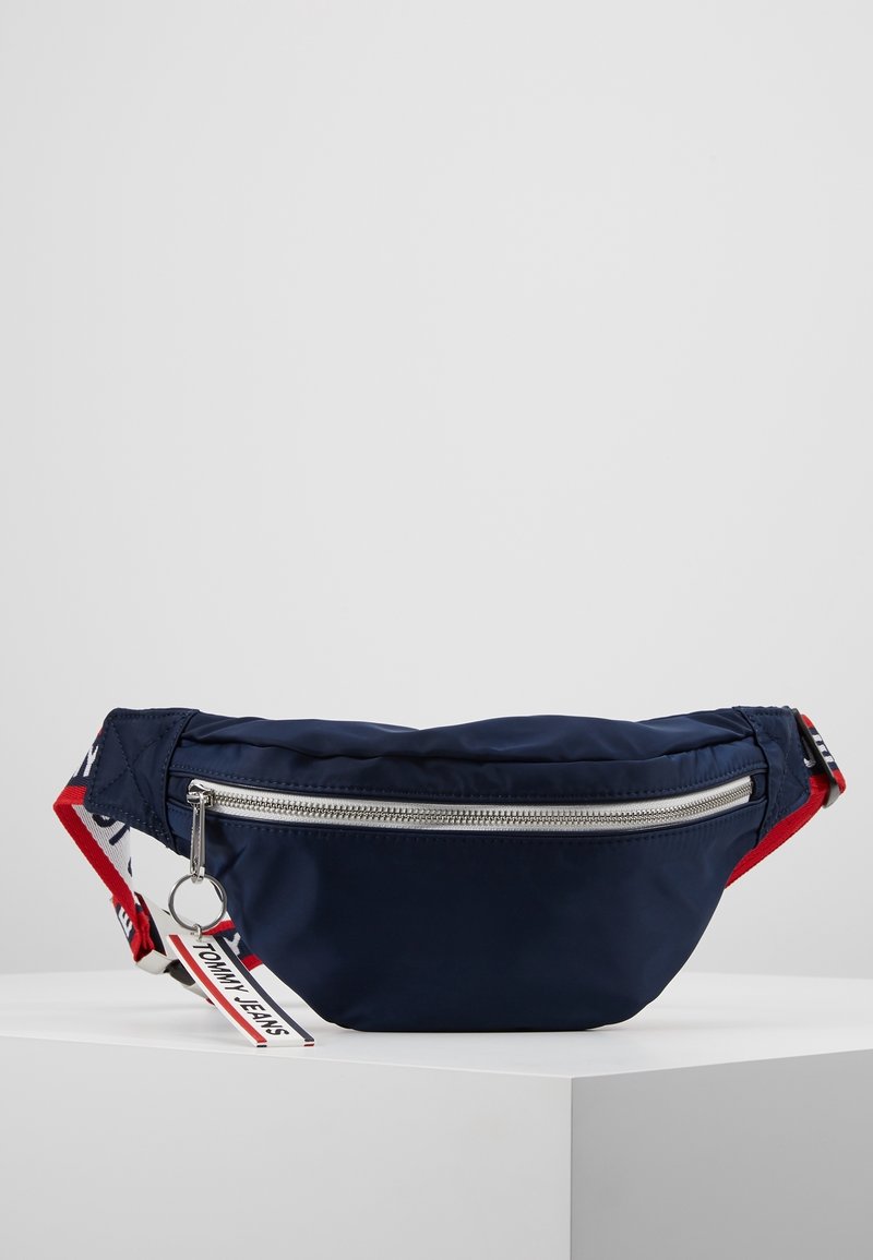 Marsupio in nylon blu navy con una cerniera argentata, una tracolla rossa e bianca e un'etichetta con il logo. Forma arrotondata con una texture liscia e una leggera lucentezza.