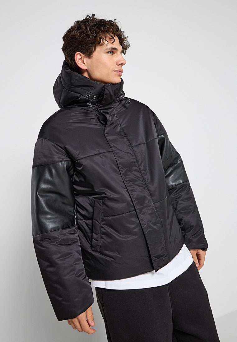 Giacca a vento nera con cappuccio, caratterizzata da una combinazione di texture opache e lucide, maniche larghe e tasche laterali. Dettagli sulle spalle.