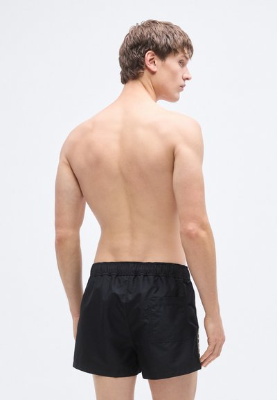 Μαύρα μαγιό σορτς με ελαστική μέση, πίσω τσέπη και απαλή, ελαφριά υφή. Το μοντέλο παρουσιάζει την πίσω όψη με ουδέτερο φόντο.