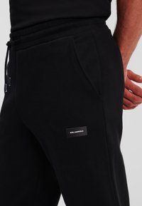 Schwarze Sweatpants mit Gummizug, Seitentaschen, weichem Material und einem kleinen Markenlabel am linken Oberschenkel.