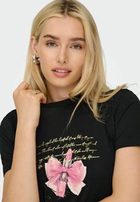 Blond kvinde iført sølvfarvede hoops øreringe og en sort skjorte med et pink læbestift og sløjfe grafik samt guld skrifttype.