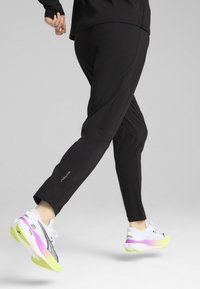 Zwarte sportbroek met taps toelopende pijpen en zijventilaties bij de enkels, gecombineerd met witte hardloopschoenen met een verloop van roze naar gele zolen.