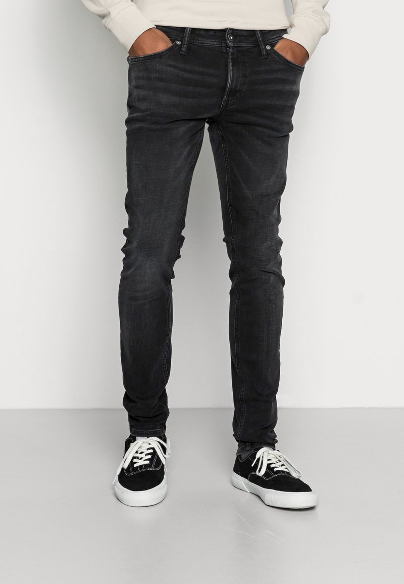 Man wearing slim-fit black jeans and black sneakers with white laces, hands in pockets, standing against a plain light background.