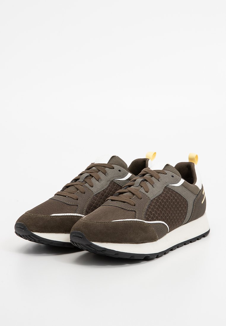 Braune Sportschuhe mit Obermaterial aus Mesh und Wildleder, runder Zehenpartie, weißen Akzenten und einer schwarzen Gummisohle mit einer gelben Zuglasche.