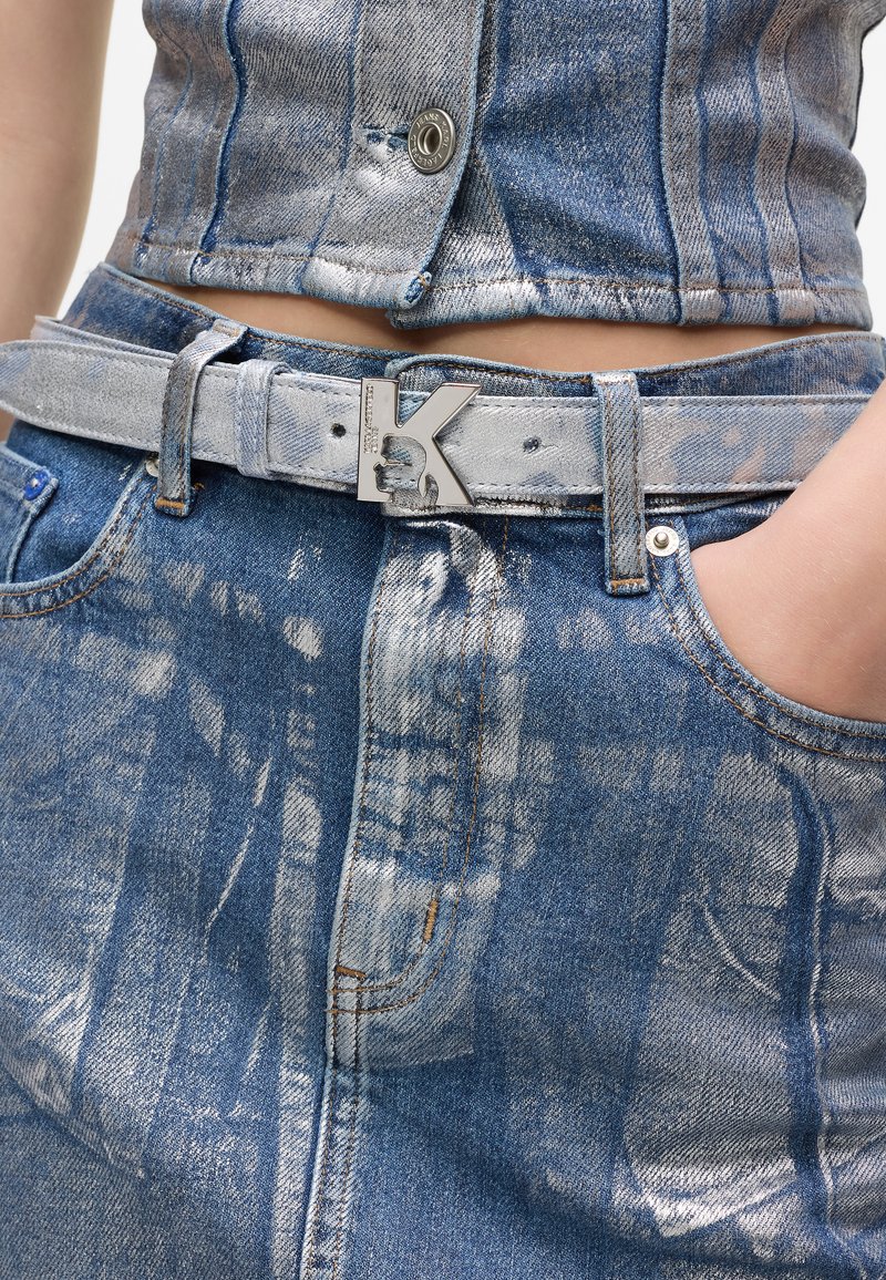 Detailní záběr na pas v modrých džínách se světlým acid-wash efektem a odpovídajícím páskem se stříbrnou sponou ve tvaru písmene "K," ruka v kapse.