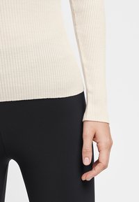 Ribbet langærmet top i off-white stof, tætsiddende design der fremhæver tekstur. Hånd hvilende på sorte leggings, som viser ærmelængden.