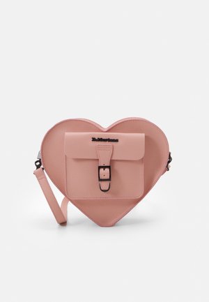 Bolso bandolera rosa con forma de corazón, con bolsillo frontal con hebilla y correa ajustable, logo de la marca Dr. Martens en la solapa del bolsillo.