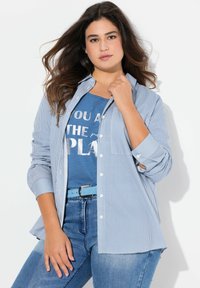 Blå och vit randig skjorta med lång ärm, i kombination med en blå grafisk t-shirt. Modell som bär blått bälte och ljusblå jeans.