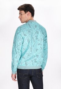 Giacca bomber turchese con un motivo astratto nero testurizzato. Presenta un colletto e polsini a coste, abbinata a jeans in denim scuro.
