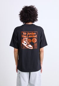 T-shirt en coton noir avec un imprimé graphique orange d'une sneaker au dos, avec du texte détaillant le matériau en cuir et la technologie anti-gravité.