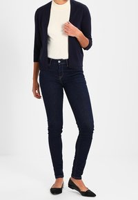 Marinblå kofta över en beige ribbad topp, kombinerad med mörka skinny jeans och svarta ballerina-skor. Koftan har trekvartslånga ärmar och öppen framsida.