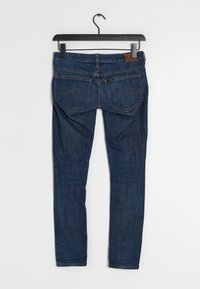 Jean en denim bleu foncé avec une coupe slim, comportant deux poches arrière, un marquage en cuir marron sur la ceinture, et des détails de couture subtils.