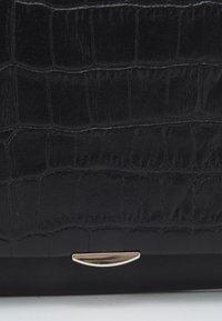 Svart handväska i konstläder med krokodilmönster, med ett guldfärgat metallfäste på botten. Strukturerad yta med glansig finish.
