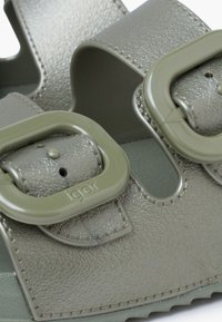 Light green sandals with two wide straps, featuring large plastic buckles. The material has a textured finish and a sturdy rubber sole.