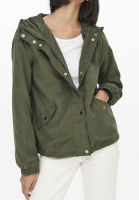 Olivengroene jas van zachte stof, met een capuchon, een rits aan de voorkant en drukknoopsluiting. Hij heeft twee zijzakken en elastische manchetten.