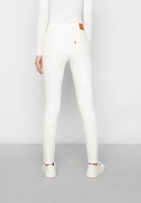 Mujer con jeans skinny blancos y zapatillas blancas con motas multicolores, de pie contra un fondo blanco liso, vista desde atrás.