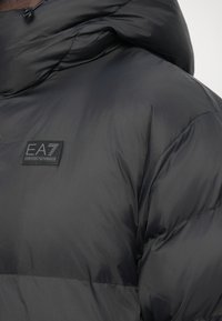 Zaprti posnetek črne puhaste jakne z visokim ovratnikom in logotipom EA7 Emporio Armani na prsih.