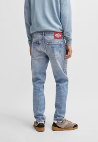 Jeans de mezclilla azul claro con un ajuste relajado, costuras prominentes y un parche de logo rojo en la parte trasera. Combinados con zapatillas beige y negras.
