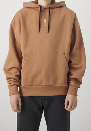 Osoba ubrana w brązową bluzę z czarnymi troczkami i przednią kieszenią, w połączeniu z czarnymi spodniami, stojąca na jednolitym tle.