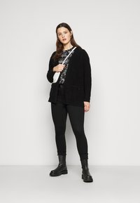 Cárdigan de punto negro con bolsillos, combinado con una camiseta gráfica negra y jeans ajustados negros. La modelo lleva botas negras de tobillo.