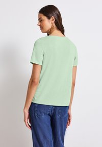 Jasnozielona koszulka z krótkim rękawem, okrągłym dekoltem i luźnym krojem. Połączona z niebieskimi dżinsami, widok z tyłu.