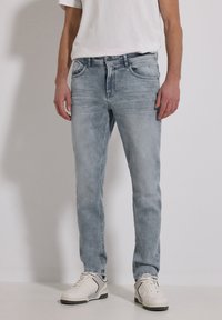 Man draagt lichtgrijze gewassen jeans, witte sneakers en een wit T-shirt, staande tegen een egale lichte achtergrond.