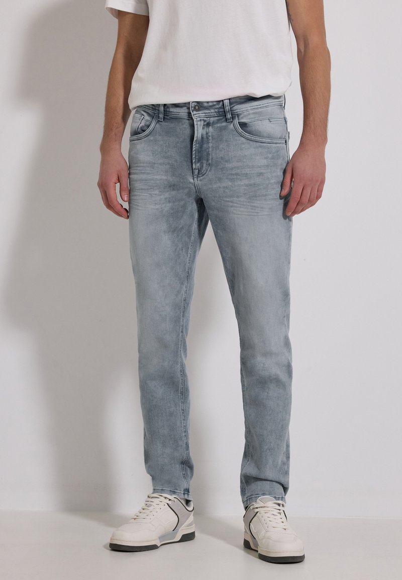 Man draagt lichtgrijze gewassen jeans, witte sneakers en een wit T-shirt, staande tegen een egale lichte achtergrond.